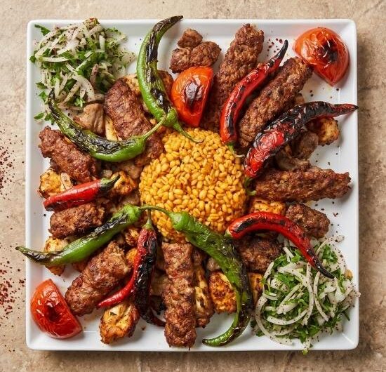 Taş zemin üzerinde kare beyaz tabakta karışık ızgara tabağının tepeden görünümü. Ortadaki bulgur pilavı, Adana kebap, tavuk şiş, köfte, közlenmiş domates ve biberlerle çevrili. Köşelerde sumaklı soğan salatası yer alıyor.